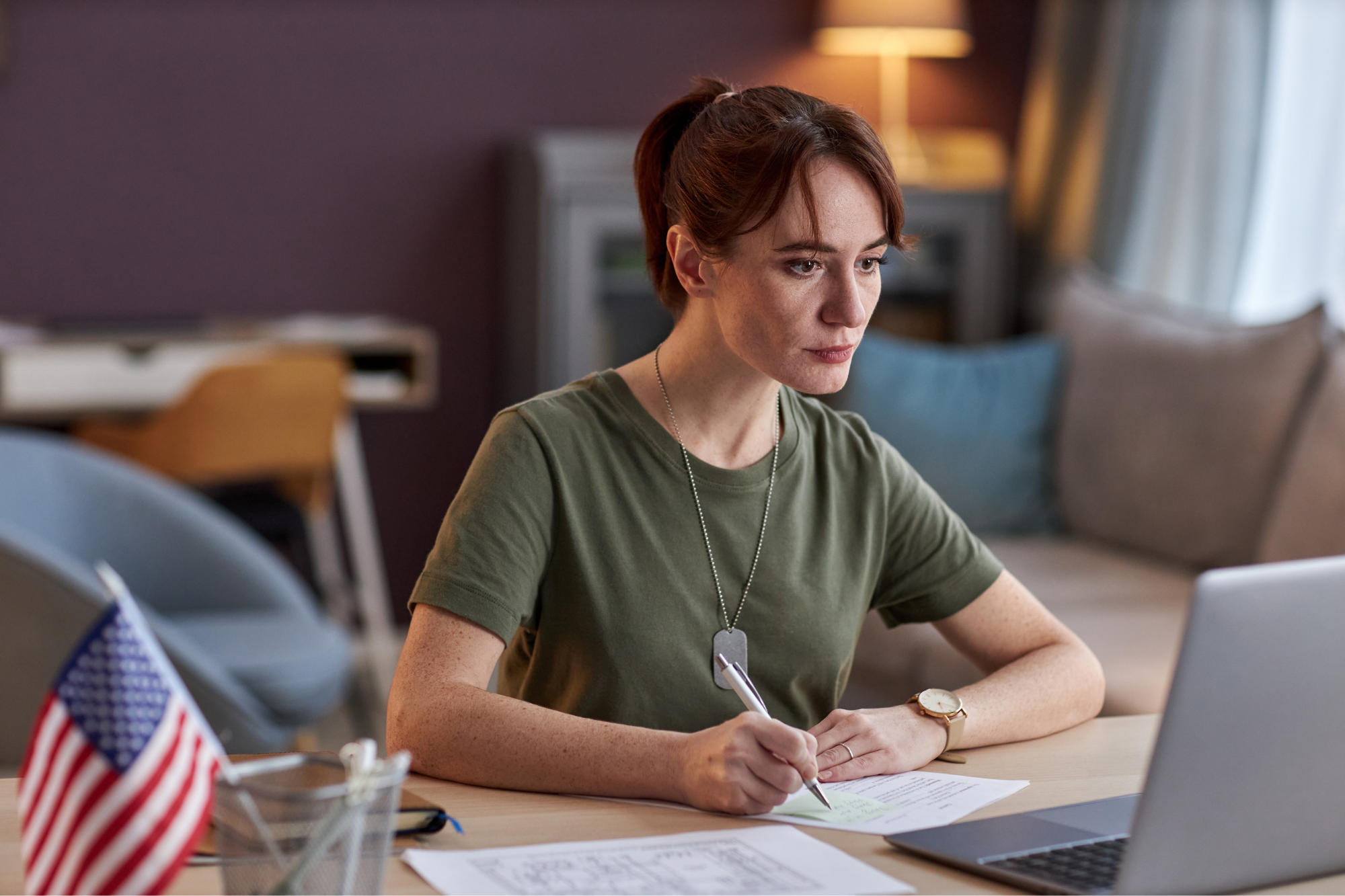 female in dark green shirt with a dog tag around her neck, sitting at a computer with an American flag on the desk